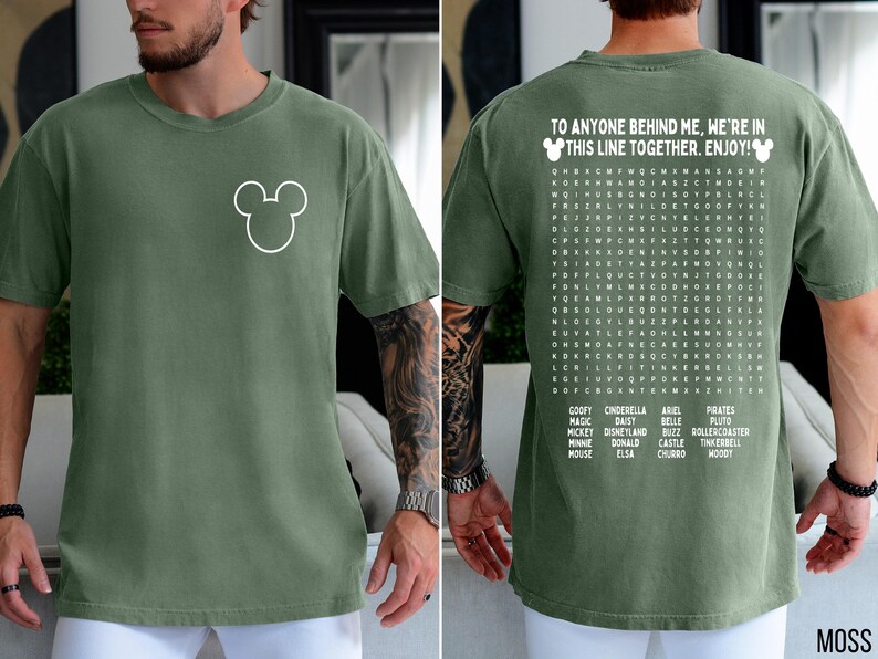 May include: Moss green t-shirt with a white Mickey Mouse outline on the front. The back of the shirt features a word search puzzle with Disney-themed words and the text "TO ANYONE BEHIND ME, WE'RE IN THIS LINE TOGETHER. ENJOY!"
