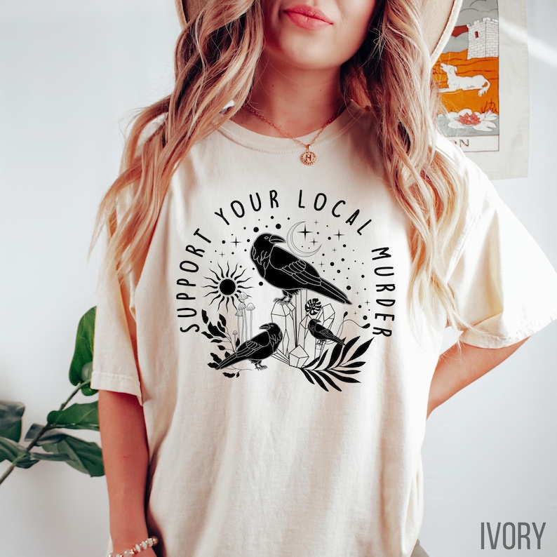 May include: Ivory-colored t-shirt featuring a black graphic of three crows perched on crystals and foliage, with a sun and moon design. The text reads "Support Your Local Murder". The shirt is a casual wear item.