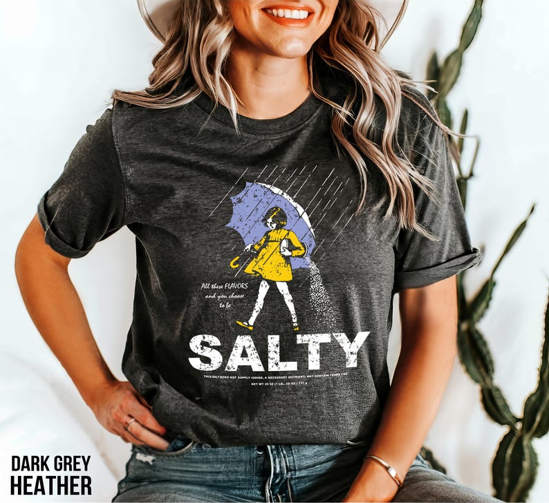 Puede incluir: Camiseta gris oscuro jaspeado con un gr&aacute;fico de una persona bajo un paraguas, con la palabra "SALTY" en blanco. La persona lleva un vestido amarillo y sostiene un paraguas azul. El texto en la camiseta dice "All them flavors and you choose to be."