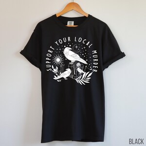 May include: Black t-shirt with a white graphic featuring three crows, a sun, moon, stars, and the text "SUPPORT YOUR LOCAL MURDER." The shirt is hanging on a wooden hanger.