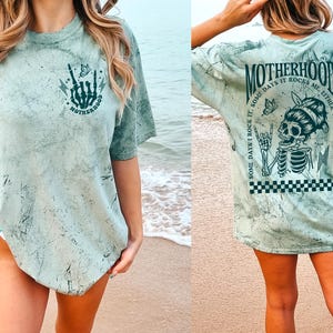 K&ouml;nnte beinhalten: Ein fern-gr&uuml;nes T-Shirt mit einem Used-Look-Design. Auf der Vorderseite befindet sich eine Skeletthand mit einem Blitz und dem Wort "MOTHERHOOD". Auf der R&uuml;ckseite ist ein Skelett mit dem Text "MOTHERHOOD, SOME DAYS I ROCK IT, SOME DAYS IT ROCKS ME, EITHER WAY WE'RE ROCKIN'" abgebildet.