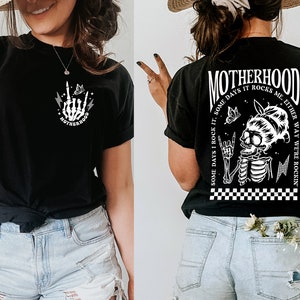 May include: Black t-shirt with a white graphic of a skeleton with a butterfly and the text "MOTHERHOOD SOME DAYS I ROCK IT, SOME DAYS IT ROCKS ME. EITHER WAY WE'RE ROCKIN'"