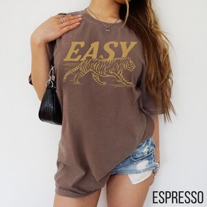 May include: Espresso-colored t-shirt with the word "EASY" in gold lettering above a gold tiger graphic. The shirt is a short-sleeved, crew neck style.