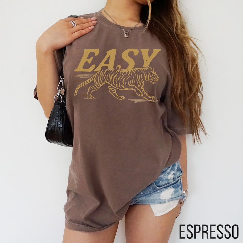May include: Espresso-colored t-shirt with the word "EASY" in gold lettering above a gold tiger graphic. The shirt is a short-sleeved, crew neck style.