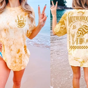 K&ouml;nnte beinhalten: Gelbes T-Shirt mit Batikmuster und dem Wort "MOTHERHOOD" sowie einer Skeletthand-Grafik auf der Vorderseite. Die R&uuml;ckseite des Shirts zeigt eine Skelettgrafik mit dem Text "SOME DAYS I ROCK IT, SOME DAYS IT ROCKS ME, EITHER WAY WE'RE ROCKIN".