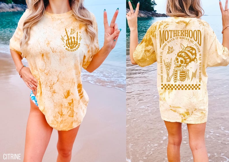 K&ouml;nnte beinhalten: Gelbes T-Shirt mit Batikmuster und dem Wort "MOTHERHOOD" sowie einer Skeletthand-Grafik auf der Vorderseite. Die R&uuml;ckseite des Shirts zeigt eine Skelettgrafik mit dem Text "SOME DAYS I ROCK IT, SOME DAYS IT ROCKS ME, EITHER WAY WE'RE ROCKIN".