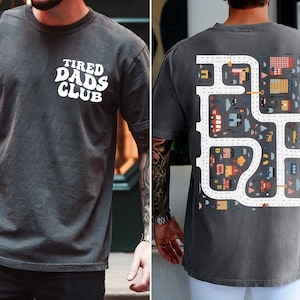 K&ouml;nnte beinhalten: Pfeffergraues T-Shirt mit dem wei&szlig;en Schriftzug "TIRED DADS CLUB" auf der Vorderseite. Auf der R&uuml;ckseite befindet sich ein farbenfrohes Stra&szlig;enkarten-Design mit Autos und Geb&auml;uden. Das Shirt hat eine lockere Passform.