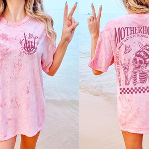 K&ouml;nnte beinhalten: Hellrosa T-Shirt mit Batikmuster und Grafik auf Vorder- und R&uuml;ckseite. Die Vorderseite zeigt eine Skeletthand mit dem Wort "MOTHERHOOD". Die R&uuml;ckseite zeigt ein Skelett mit Kopftuch, die Worte "MOTHERHOOD" und den Satz "SOME DAYS I ROCK IT, SOME DAYS IT ROCKS ME, EITHER WAY WE'RE ROCKIN".