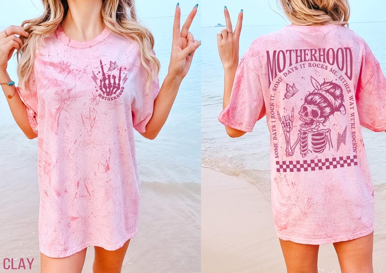 K&ouml;nnte beinhalten: Hellrosa T-Shirt mit Batikmuster und Grafik auf Vorder- und R&uuml;ckseite. Die Vorderseite zeigt eine Skeletthand mit dem Wort "MOTHERHOOD". Die R&uuml;ckseite zeigt ein Skelett mit Kopftuch, die Worte "MOTHERHOOD" und den Satz "SOME DAYS I ROCK IT, SOME DAYS IT ROCKS ME, EITHER WAY WE'RE ROCKIN".
