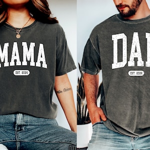 May include: Two gray t-shirts with white text. The shirt on the left says "MAMA EST. 2024" and the shirt on the right says "DAD EST. 2024".
