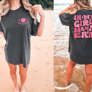 May include: A charcoal grey short-sleeve t-shirt featuring a small pink smiley face graphic on the front left chest with the text "GIRL MAMA ERA". The back displays a large pink graphic with the text "IN MY GIRL MAMA ERA" and smiley faces.