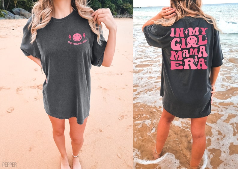 May include: A charcoal grey short-sleeve t-shirt featuring a small pink smiley face graphic on the front left chest with the text "GIRL MAMA ERA". The back displays a large pink graphic with the text "IN MY GIRL MAMA ERA" and smiley faces.