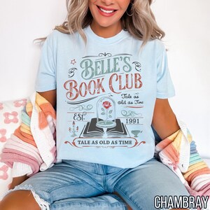 May include: Light blue t-shirt with the text "Belle's Book Club" in a vintage design. The graphic includes a rose in a glass dome, the words "Tale as old as Time," and the year 1991. The shirt is made of soft material.
