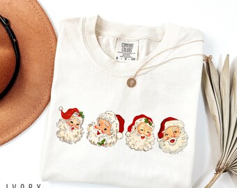 May include: Ivory t-shirt with a vintage-style graphic of four Santa Claus faces. The shirt has a crew neck and short sleeves. The text "COMFORT COLORS" is visible on the tag.