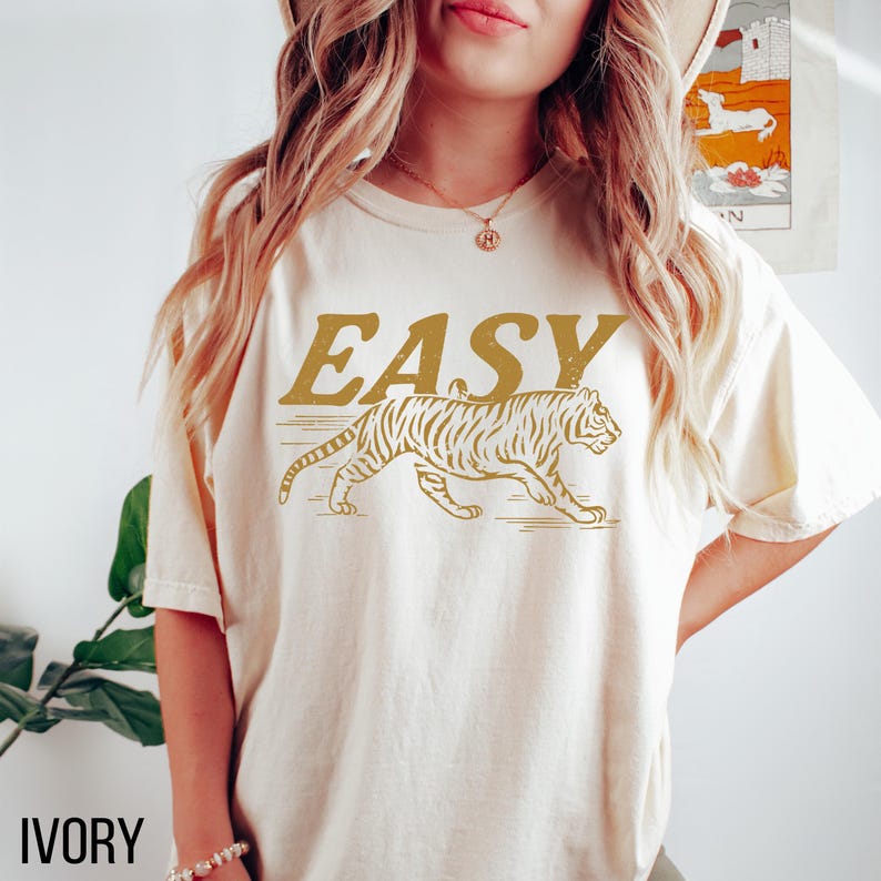 May include: Ivory-colored t-shirt with the word "EASY" in a gold, vintage font above a gold tiger graphic. The shirt is a relaxed fit and has short sleeves. The word "IVORY" is printed in a black font at the bottom.