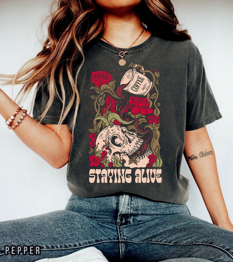 May include: A dark grey t-shirt with a graphic of a skull with red roses and a coffee cup pouring coffee into the skull. The text "STAYING ALIVE" is printed below the graphic.