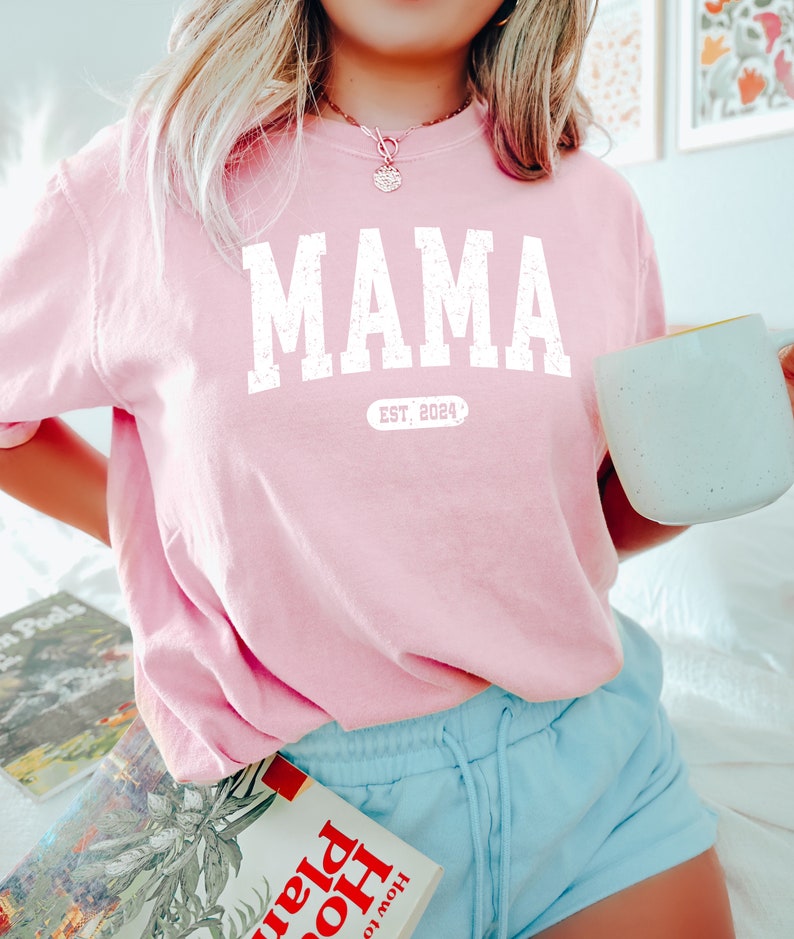 May include: Pink t-shirt with the word "MAMA" in white with a distressed font and the text "EST. 2024" underneath.