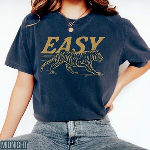 May include: Navy blue t-shirt with the word "EASY" in gold lettering above a gold tiger illustration. The tiger is walking to the right. The shirt is a short-sleeved, crew neck style.