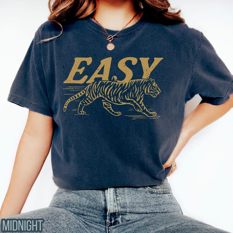 May include: Navy blue t-shirt with the word "EASY" in gold lettering above a gold tiger illustration. The tiger is walking to the right. The shirt is a short-sleeved, crew neck style.