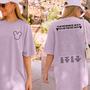 May include: Orchid-colored oversized t-shirt featuring a black Mickey Mouse outline on the front. The back of the shirt has a word search puzzle with Disney-related words and the text "TO ANYONE BEHIND ME, WE'RE IN THIS LINE TOGETHER. ENJOY!"