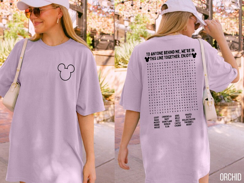 May include: Orchid-colored oversized t-shirt featuring a black Mickey Mouse outline on the front. The back of the shirt has a word search puzzle with Disney-related words and the text "TO ANYONE BEHIND ME, WE'RE IN THIS LINE TOGETHER. ENJOY!"