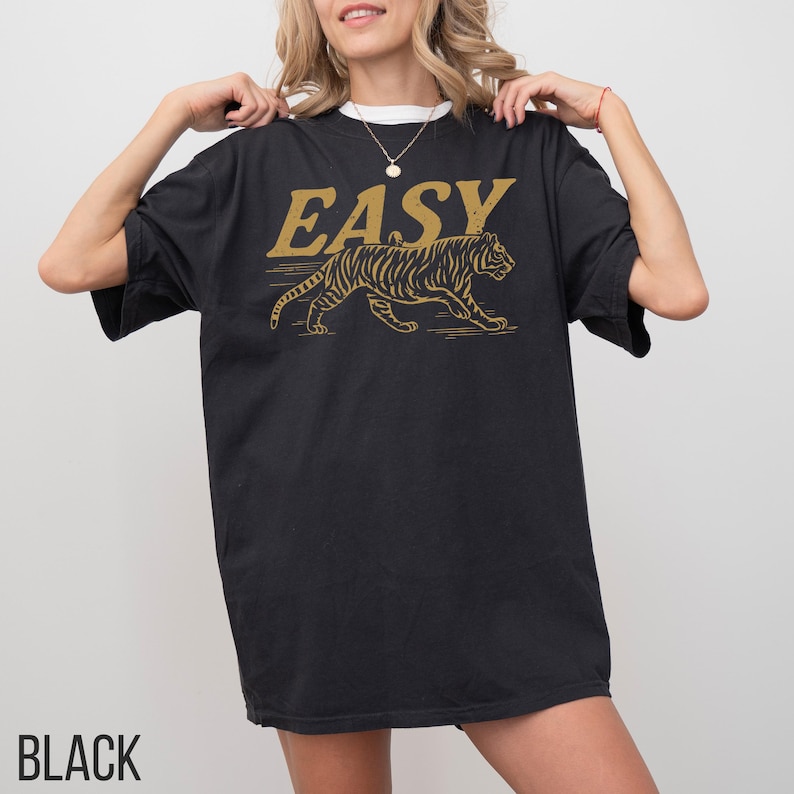 May include: Black t-shirt with the word "EASY" in gold lettering above a gold tiger graphic. The shirt has short sleeves and a relaxed fit. The word "BLACK" is printed at the bottom of the shirt.