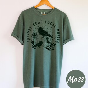 May include: Moss green t-shirt with a black graphic featuring three crows, a sun, moon, and the text "Support Your Local Murder". The shirt is hanging on a wooden hanger.