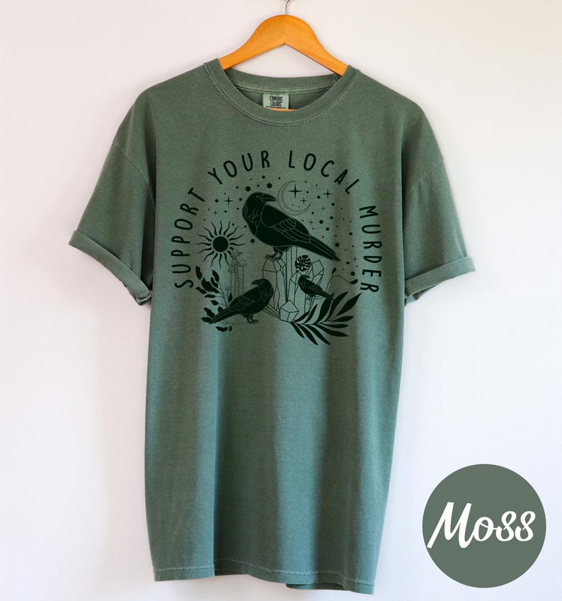 May include: Moss green t-shirt with a black graphic featuring three crows, a sun, moon, and the text "Support Your Local Murder". The shirt is hanging on a wooden hanger.