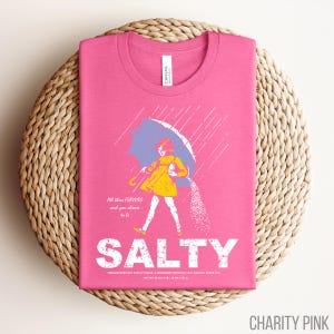 Puede incluir: Camiseta rosa con un gr&aacute;fico de una persona bajo un paraguas, vertiendo sal. La palabra "SALTY" est&aacute; impresa en letras blancas grandes debajo de la ilustraci&oacute;n. El texto "All these flavors and you choose to be" est&aacute; arriba.