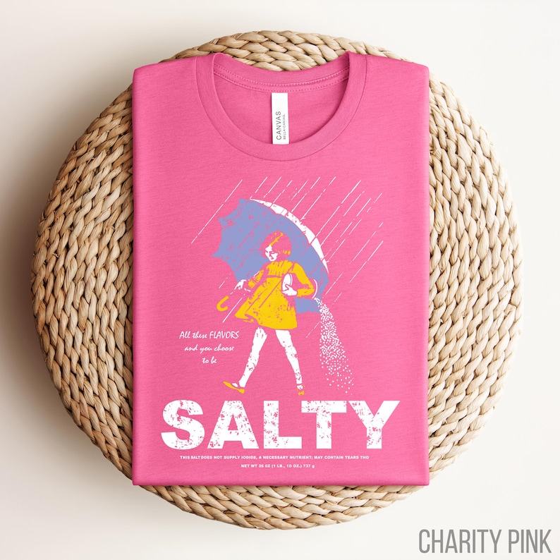 Puede incluir: Camiseta rosa con un gr&aacute;fico de una persona bajo un paraguas, vertiendo sal. La palabra "SALTY" est&aacute; impresa en letras blancas grandes debajo de la ilustraci&oacute;n. El texto "All these flavors and you choose to be" est&aacute; arriba.