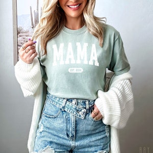 May include: A light green t-shirt with the word "MAMA" in white block letters and the text "EST. 2024" underneath. The shirt is being worn by a person with long blonde hair and is paired with denim shorts.