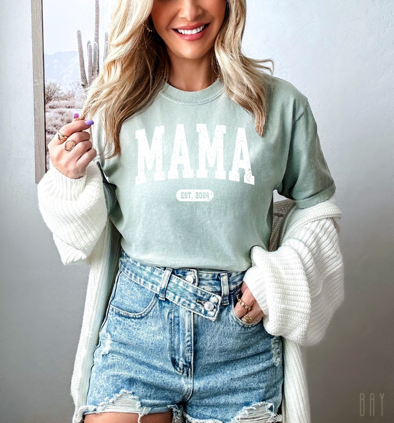 May include: A light green t-shirt with the word "MAMA" in white block letters and the text "EST. 2024" underneath. The shirt is being worn by a person with long blonde hair and is paired with denim shorts.