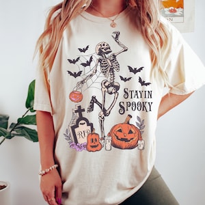May include: A beige t-shirt with a graphic of a skeleton holding a jack-o-lantern and dancing on a spider web. The graphic also includes a tombstone, pumpkins, and bats. The text "Stayin Spooky" is printed on the shirt.