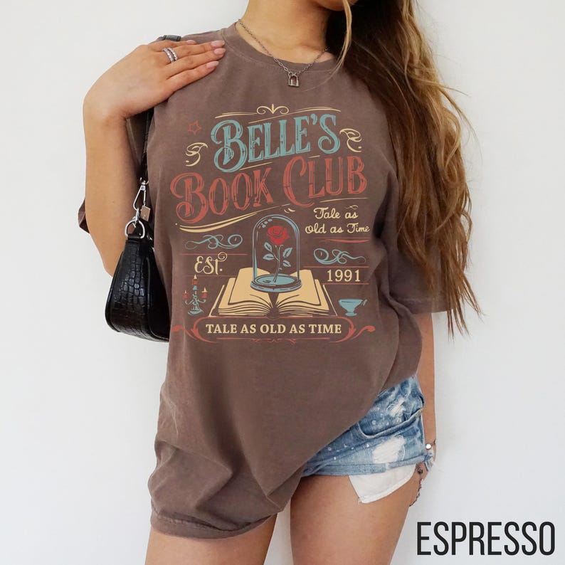 May include: Espresso-colored t-shirt with the text "Belle's Book Club" in a vintage design. The design includes a rose in a glass case, the words "Tale as Old as Time," and the year 1991.
