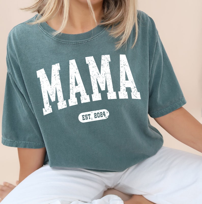 May include: A teal blue t-shirt with the word "MAMA" in white distressed lettering and the text "EST. 2024" in a white oval.