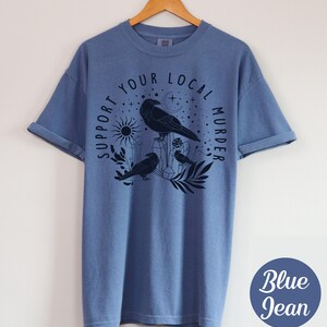 May include: Blue jean-colored t-shirt with a graphic of three crows, crystals, a sun, and the text "Support Your Local Murder." The design is in black. The shirt is hanging on a wooden hanger.