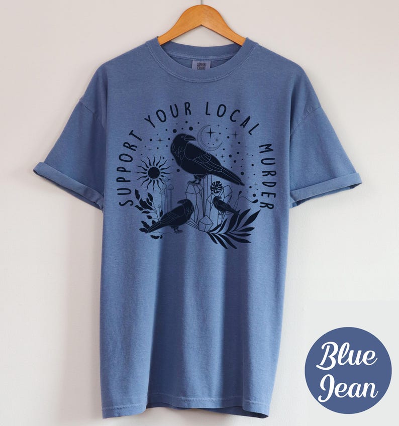 May include: Blue jean-colored t-shirt with a graphic of three crows, crystals, a sun, and the text "Support Your Local Murder." The design is in black. The shirt is hanging on a wooden hanger.
