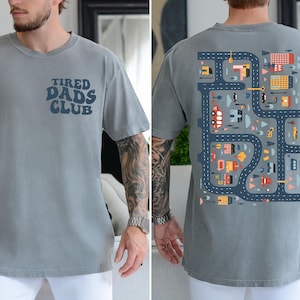 K&ouml;nnte beinhalten: Graues Kurzarm-T-Shirt mit dem Text "TIRED DADS CLUB" auf der Vorderseite. Die R&uuml;ckseite zeigt ein farbenfrohes Stra&szlig;enkarten-Design mit Fahrzeugen und Geb&auml;uden. Das Shirt ist aus weichem Material.