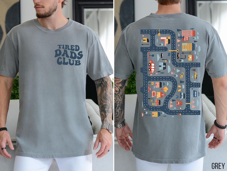 K&ouml;nnte beinhalten: Graues Kurzarm-T-Shirt mit dem Text "TIRED DADS CLUB" auf der Vorderseite. Die R&uuml;ckseite zeigt ein farbenfrohes Stra&szlig;enkarten-Design mit Fahrzeugen und Geb&auml;uden. Das Shirt ist aus weichem Material.