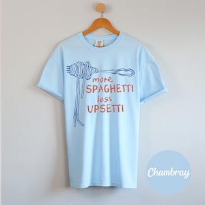 May include: Light blue t-shirt with a blue and red graphic of spaghetti on a fork. The text on the shirt reads "more spaghetti less upsetti".