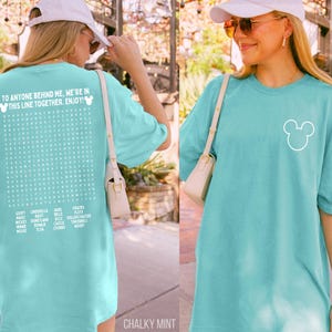 May include: A chalky mint-colored t-shirt with a white Mickey Mouse outline on the front. The back of the shirt features a word search with Disney-related words and the text "TO ANYONE BEHIND ME, WE'RE IN THIS LINE TOGETHER. ENJOY!"