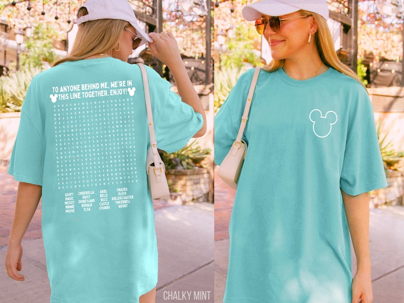 May include: A chalky mint-colored t-shirt with a white Mickey Mouse outline on the front. The back of the shirt features a word search with Disney-related words and the text "TO ANYONE BEHIND ME, WE'RE IN THIS LINE TOGETHER. ENJOY!"