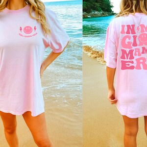 May include: A light pink t-shirt featuring a small pink smiley face graphic with lightning bolts and the text "GIRL MAMA ERA" on the front chest, and a large version of the same graphic and text printed across the back of the garment.