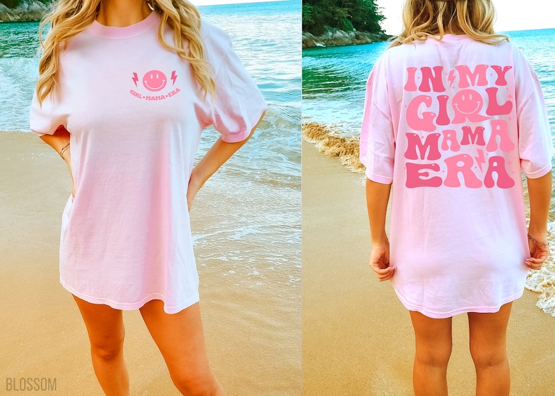 May include: A light pink t-shirt featuring a small pink smiley face graphic with lightning bolts and the text "GIRL MAMA ERA" on the front chest, and a large version of the same graphic and text printed across the back of the garment.