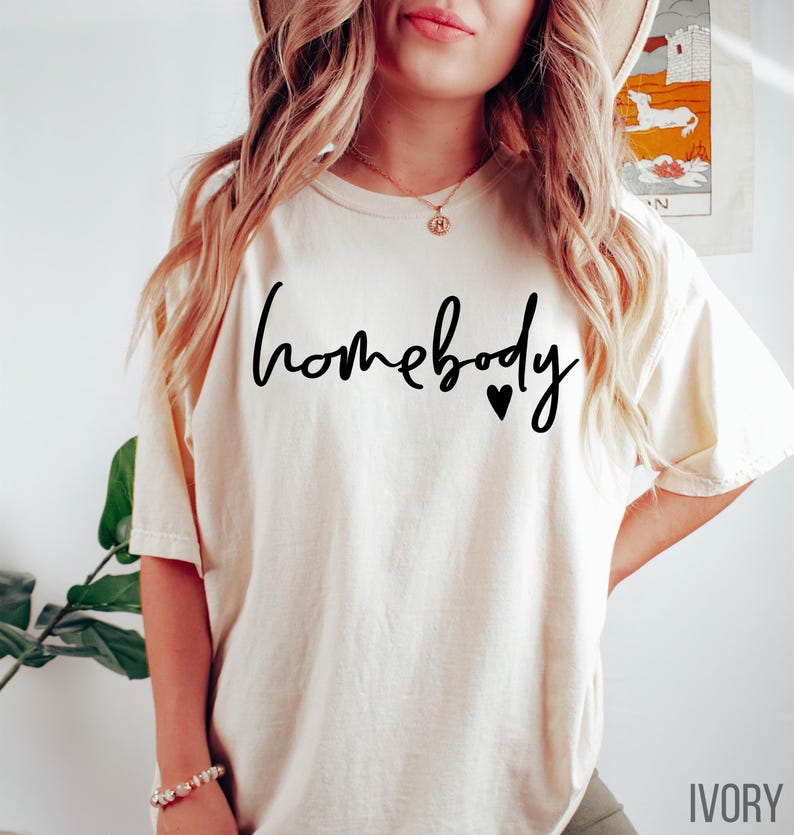 Puede incluir: Una camiseta blanca con el texto "homebody" en escritura negra con dos peque&ntilde;os corazones negros. La camiseta se muestra en una modelo y el color se etiqueta como "marfil".