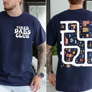K&ouml;nnte beinhalten: Marineblaues T-Shirt mit dem Text "TIRED DADS CLUB" auf der Vorderseite. Die R&uuml;ckseite des Shirts zeigt ein farbenfrohes Stra&szlig;enkarten-Design mit Autos, Geb&auml;uden und B&auml;umen. Das Shirt ist ein bequemer, l&auml;ssiger Stil.