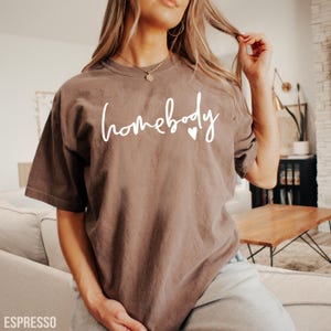 Puede incluir: Una camiseta marr&oacute;n con el texto "homebody" en letras blancas y dos peque&ntilde;os corazones blancos.