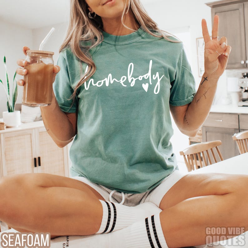 Puede incluir: Una camiseta azul turquesa con el texto "homebody" en letras blancas con un peque&ntilde;o coraz&oacute;n blanco. La camiseta la lleva puesta una persona sentada en una silla.