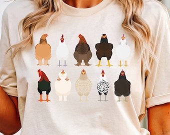 May include: An ivory short-sleeve t-shirt featuring a graphic print of ten different chicken breeds arranged in two rows. The chickens are depicted in various colors including brown, white, black, and speckled patterns, each with distinct plumage.