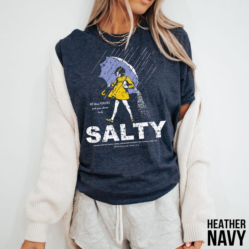 Puede incluir: Camiseta azul marino jaspeado con un gr&aacute;fico de una persona caminando bajo la lluvia con un paraguas, con la palabra "SALTY" en letras blancas grandes. La persona lleva un abrigo amarillo. El texto de la camiseta dice "All these flavors and you choose to be".